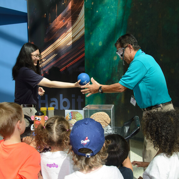 cation team member and Volunteer demonstrate science concept at a cart by passing each other a small blue rubber ball while group of kids watch