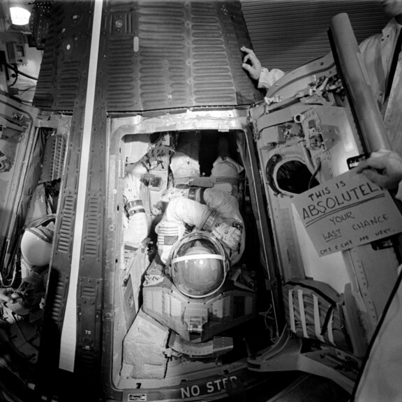 Conrad (left) and Gordon (center) are inserted into their couches aboard Gemini XI on launch morning. A sign reads 'This is absolutely your last chance. CM3 & CM4 are next.'