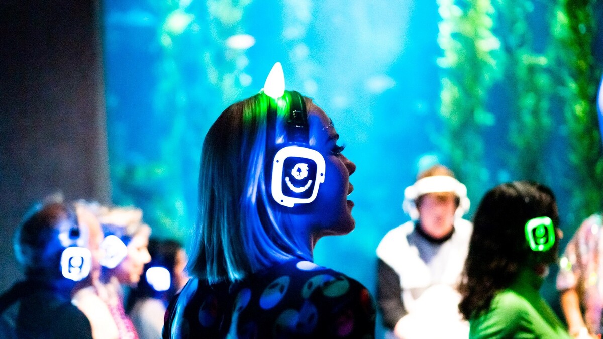 Person wearing neon lit headphones and dancing to silent disco while DJ mixes in front of Ecosystems Kelp Tank
