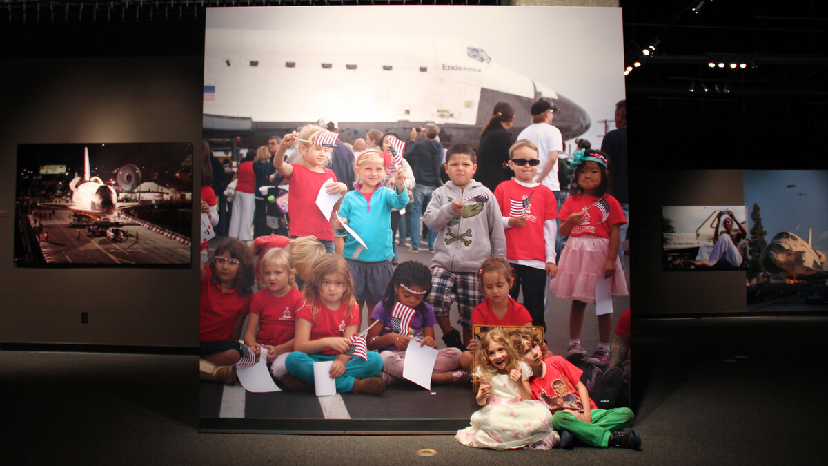 Two kids sit in front of a large mural of Endeavour, sitting in a parking lot with a class of schoolkids