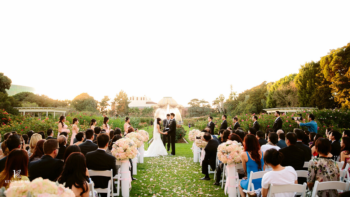 Wedding guests sit in white folding chairs overlooking a wedding ceremony in the Exposition Park Rose Garden