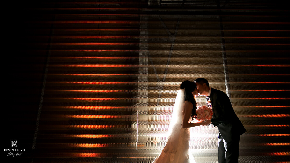 A bride and groom kiss at dusk in front of corrugated metal lit in orange