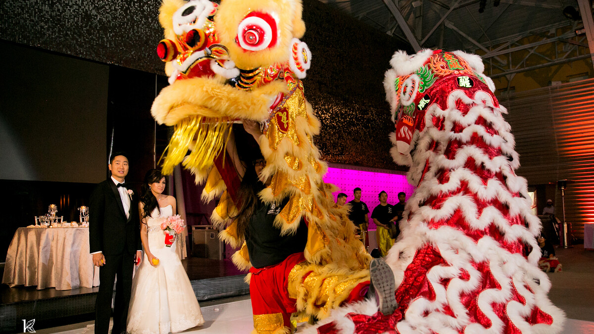 A bride and groom watch as red and yellow traditional lion dancers perform on the dancefloor