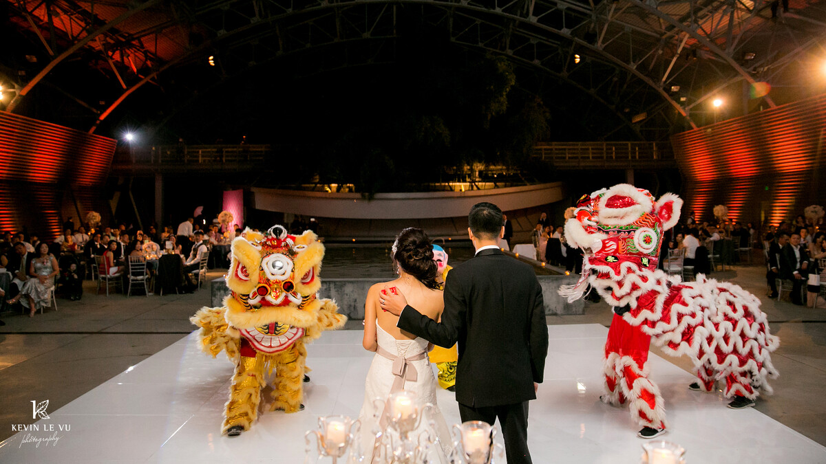 A bride and groom watch as red and yellow traditional lion dancers perform on the dancefloor in the Wallis Annenberg Building: Big Lab