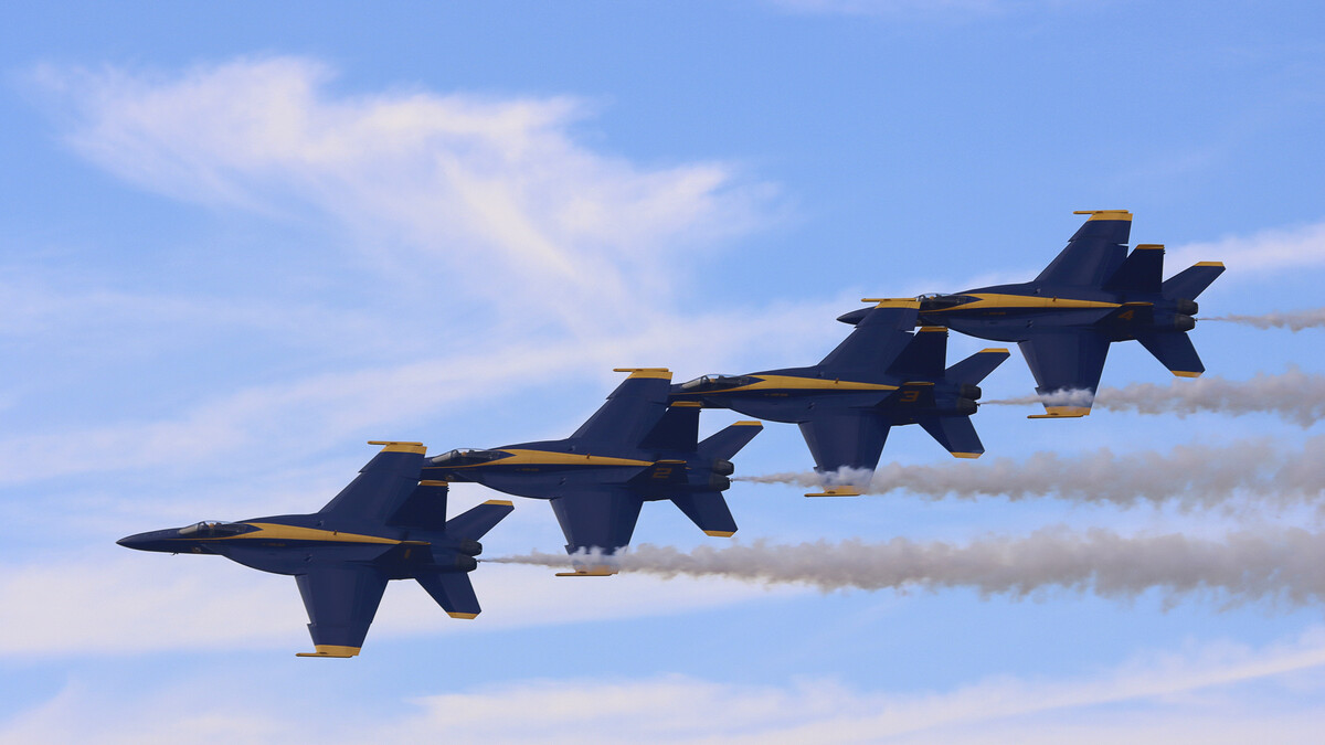 The Blue Angels fly in formation through a blue sky with wispy clouds, leaving behind a trail from their planes.