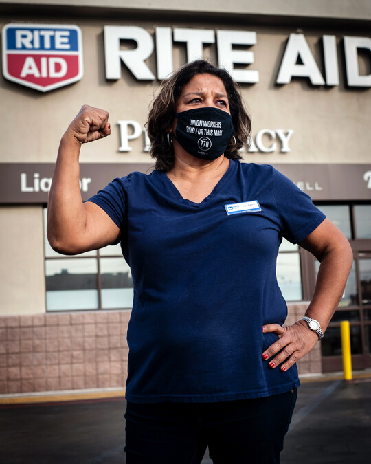 Laura Orantes, retail health clerk, stands with her arm raised in solidarity in front of a Rite Aid pharmacy