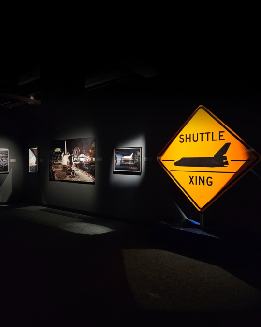 An orange traffic sign that says "SHUTTLE XING" next to wall mounted photos inside the Work in Progress gallery